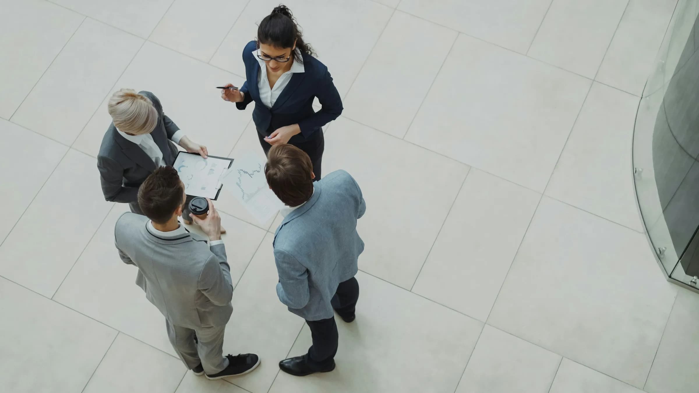 Four colleagues in business clothes, seen from above, standing together over a clipboard and a printed chart in a bright office lobby.